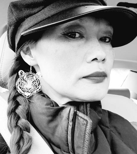 A black and white portrait of me in a car, looking towards the camera. I am wearing a black newsboy cap and a large tiger head earring. My hair is braided, I'm wearing a black vest jacket, with a seatbelt on. I have a serious expression, with defined eye make-up and dark lipstick.