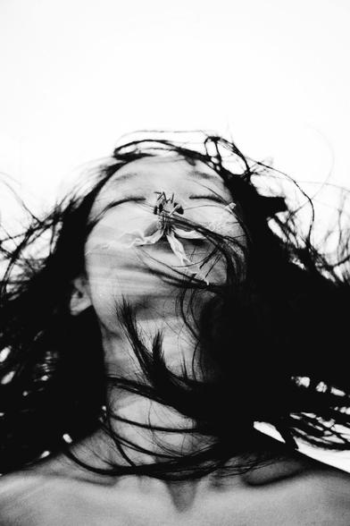 Black and white artistic photograph of an Asian woman's face, with head tilted back and eyes closed, and a flower placed over mouth and nose, with flowing dark hair.