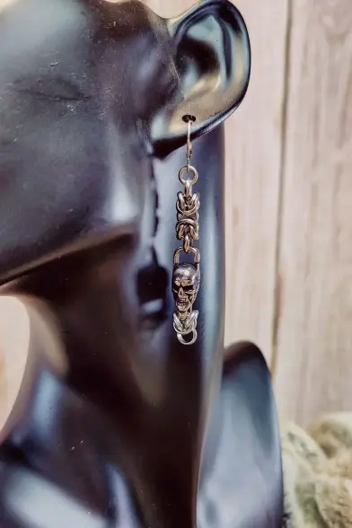 Boucle d'oreille en maille byzantine et intégrant un petit crâne en acier inoxydable. La boucle est installée sur un présentoir pour montrer le rendu quand elle est portée.