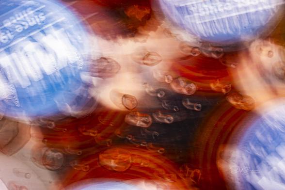 Ein abstraktes Bild mit verschwommenen blauen Flaschenverschlüssen vor einem Hintergrund aus leuchtenden rötlichen Tönen. Die Komposition enthält Blasen und ein Gefühl von Bewegung, wodurch ein dynamischer visueller Effekt entsteht.