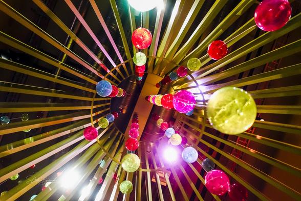 Eine künstlerische Ansicht mit Blick auf eine kreisförmige Deckenstruktur, die mit bunten Glühbirnen in Rot-, Grün-, Blau- und Gelbtönen verziert ist, umgeben von linearen Metallbalken. Die Beleuchtung schafft eine lebendige und fröhliche Atmosphäre.