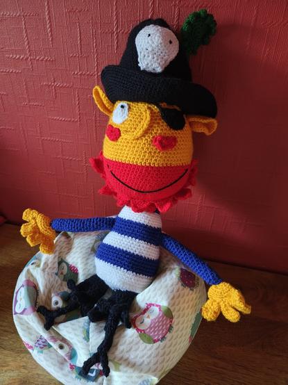 The crochet version of the pirate in the previous image sitting on a small owl beanbag next to a red wall. He is looking at the camera and smiling.