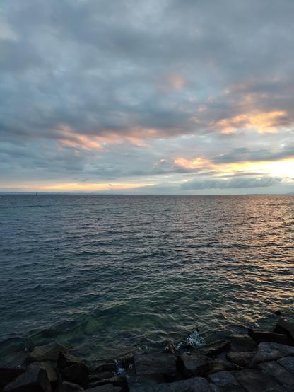 Blick von Romanshorn Hafen Richtung Osten über den Bodensee, Morgenhimmel mit Wolken, rechts drückt die Sonne durch, färbt Wolken orange und rosa, das andere Ufer ist im Dunst knapp zu sehen, links die Leuchtturmdalbe, bewegte Wasseroberfläche, blau-silbriges Wasser, im Vordergrund Steinblöcke der Uferbefestigung