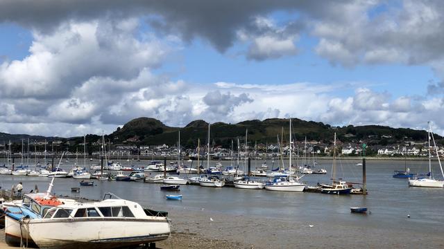 A small harbour contains numerous moored boats, including sailboats and motorboats, with a few resting on the muddy shore in the foreground. Calm waters reflect the vessels, and the tide appears low, leaving parts of the shoreline exposed. Across the water, a cluster of houses stretches along the base of rolling green hills, with scattered trees and shrubbery dotting the slopes. The sky is filled with a mix of dark and white clouds, allowing patches of blue to break through, casting alternating shadows and light over the scene.
