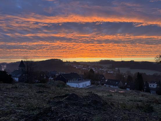 Ein kleines Dorf in hügeliger Landschaft bei Sonnenaufgang: dunkle Häuser im Vordergrund, dahinter Felder und Wälder, über allem ein dramatischer Himmel in leuchtenden Orange‑, Rot‑ und Blautönen.