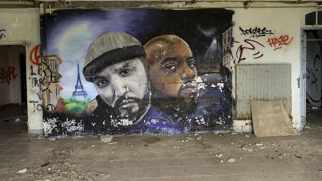 Hier sieht man ein großes Graffiti an einer alten Wand, auf denen zwei Personen zu sehen sind. Links von ihnen ist der kleine Eiffelturm zu sehen. Das Graffiti ist sehr farbenfroh.