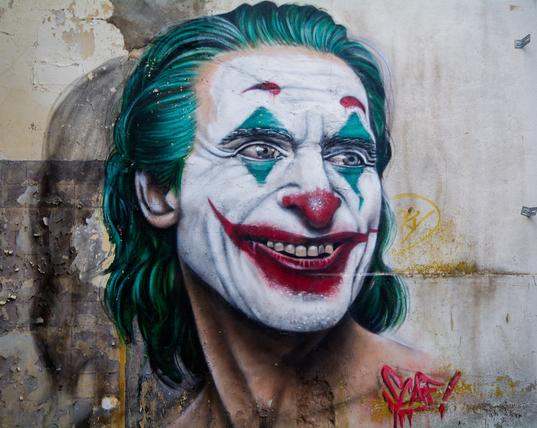 Das Bild zeigt ein Graffito des Jokers, der eine realistische Darstellung der Figur ist. Er hat das typische grüne Haar, die weiße Gesichtsbemalung, die rote Nase und den roten Lippenstift, die ihm ein breites, unheimliches Lächeln verleihen. Das Graffiti scheint auf eine abgenutzte oder beschädigte Wand gesprüht worden zu sein, was dem Bild einen urbanen und düsteren Touch verleiht. Am Hals des Jokers befindet sich eine rote Kritzelei, die wie ein Tag oder eine Signatur aussieht.