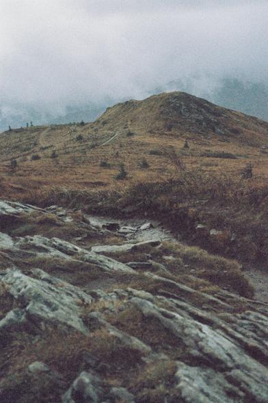 A muddy path leading through heathland and dry grass, with mist-covered mountains and breaking clouds in the background.