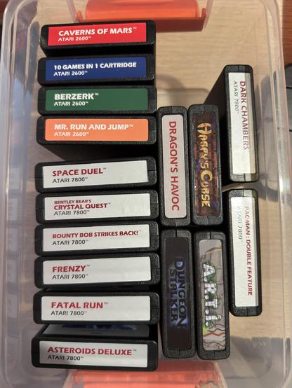 Una colección de cartuchos de juego Atari antiguos expuestos en un contenedor transparente, con títulos como "Caverns of Mars", "Berzerk", "Dragon's Havoc" y "Asteroids Deluxe". Los cartuchos están etiquetados con sus respectivos títulos de juego