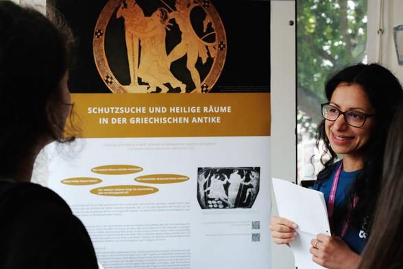 In einem Zelt steht eine Posterwand. Daran hängt ein Poster. Es zeigt altgriechische Motive und die Überschrift "Schutzsuche und Heilige Räume in der griechischen Antike". Rechts neben dem Poster steht eine Frau. Sie hält Papiere in den Händen und spricht mit einer Person, die links schräg vor dem Poster steht.
