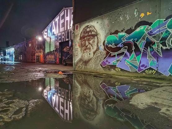 Mehrere große Graffiti an einer Betonwand spiegeln sich nachts in einer großen Pfütze. Im Hintergrund die beleuchtete Skatehalle Berlin.