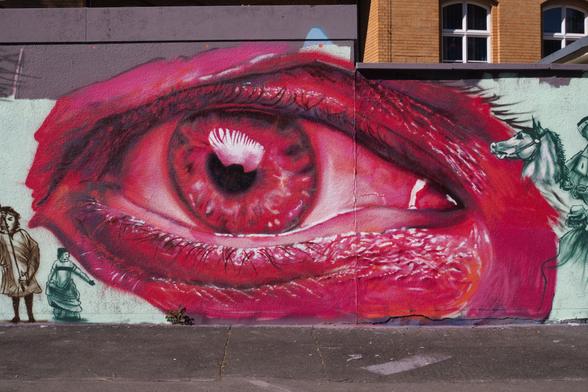 Das Bild zeigt ein großes, sehr detailreiches Graffiti eines pink-roten Auges zu sehen. Das Auge nimmt fast die gesamte Breite der Wand ein und wirkt durch seine kräftigen Farben und die feinen Linien sehr lebendig. Links und rechts neben dem Auge sind kleinere, skizzenhafte Figuren zu erkennen – links zwei Menschen in einfacher Darstellung, rechts ein Reiter auf einem Pferd. Der Boden vor der Wand ist grau und wirkt wie ein normaler Gehweg.