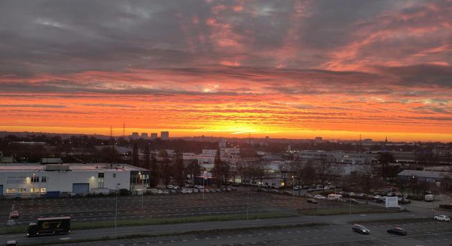 Roter Himmel, erste Sonnenstrahlen leuchten von unten an die Wolkendecke. Im Vordergrund ein fast leerer großer Parkplatz.
