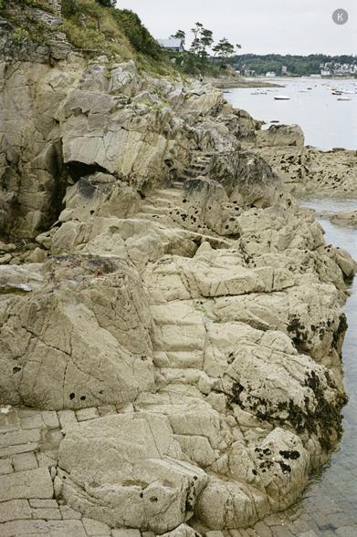Küstenfelsen mit natürlichen Stufen am Rand eines klaren, flachen Meeres; im Hintergrund liegen kleine Boote in einer ruhigen Bucht unter bewölktem Himmel.