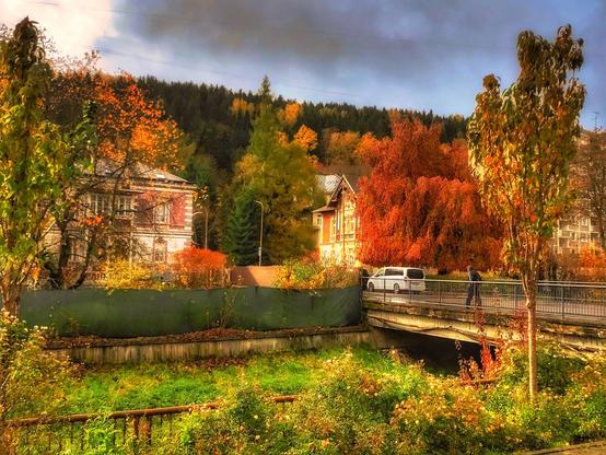 Rechts eine Brücke über einen kleinen Fluss. Dahinter Häuser, die vom herbstlich roten Laub der Bäume teilweise verdeckt sind. Der Himmel teilweise blau mit dunklen Wolken.