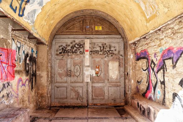 A weathered, arched wooden doorway set into a crumbling, yellowish stucco wall, heavily adorned with graffiti. The door itself is aged, with peeling paint and graffiti scrawled across its panels. Above the door, the number '29' is visible on a small blue plaque, and a yellowing sign is affixed nearby. The surrounding walls are covered in vibrant and abstract graffiti, adding a stark contrast to the dilapidated, rustic charm of the narrow passageway.