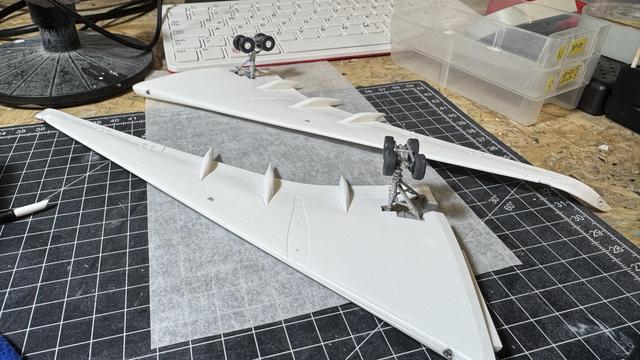 Two white model airplane wings placed upside down on a workbench with visible landing gears attached, surrounded by a cutting mat and various tools.