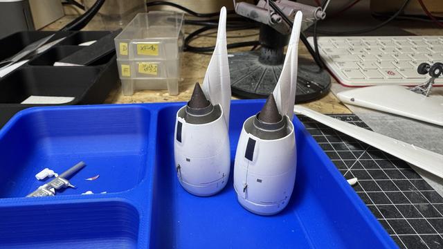 Two model airplane jet engines with attached vertical stabilizers placed in a blue plastic tray on a workbench, surrounded by modeling tools and materials.