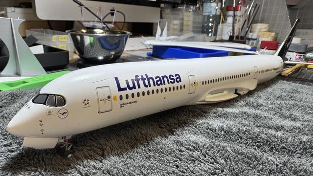 Close-up of the front section of a detailed Lufthansa Airbus A350-900 model airplane, resting on a textured surface with a computer keyboard and workspace in the background.
