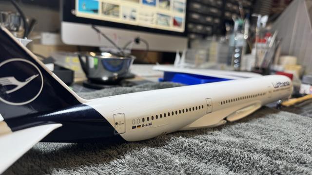 Side view of the Lufthansa Airbus A350-900 model airplane showing the fuselage and tail fin with the Lufthansa logo, positioned on a textured surface with tools and workspace in the background. 