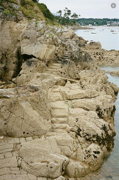 Küstenfelsen mit natürlichen Stufen am Rand eines klaren, flachen Meeres; im Hintergrund liegen kleine Boote in einer ruhigen Bucht unter bewölktem Himmel.