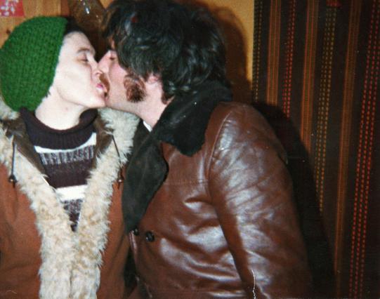A scan of a late 1970s colour photo in the living room of a house, late at night as the curtains are closed and the people dressed for outside as if they're about to leave or have just come in from the cold. The walls are pale orange at the back and pine-panelled on the left where a dimpled pint glass can be seen hanging from a shelf hook. Curtains on the right side are dark brown and patterned with a series of thin stripes and small vertical dots in oranges, yellows, and lighter browns. The main focus is just left of view where a couple are leaning in to kiss one another, deep shadows on the wall behind from the camera flash. The woman on the left where a black and white hooped jumper over which she has a brown coat with white fake fur trim. She wears a green, bobbly, knitted beret-style hat. The main has a brown leather coat on with a large, black collar. He's dark-haired with a moustache and sideburns. It's a very 70s smooch.