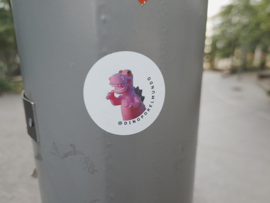 A small circular white sticker on a gray pole features a pink, smiling, cartoon dinosaur giving a thumbs-up. Text around the bottom edge reads "@DINOPORELMUNDO".