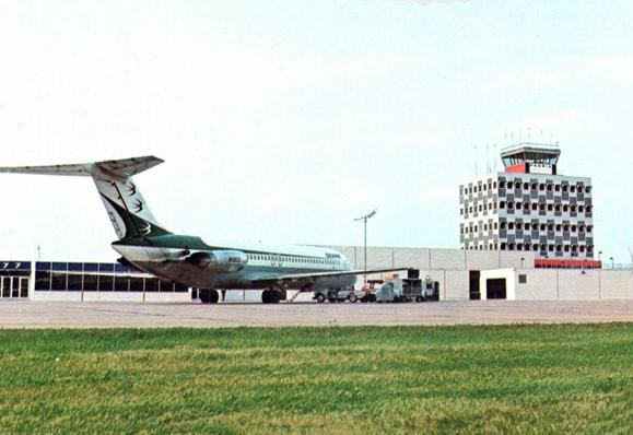 he postcard captures an Ozark Air Lines McDonnell Douglas DC-9 parked on the ramp at Greater Peoria Airport in Illinois, likely in the early 1970s. The aircraft wears Ozark’s iconic green-and-silver livery featuring the stylized “Three Swallows” logo on the tail — one of the most recognizable regional airline identities of the era. The DC-9, a short-to-medium range twinjet, was the backbone of Ozark’s fleet and perfectly suited for linking Midwestern cities like Peoria with major hubs such as St. Louis and Chicago.

Behind the aircraft stands the mid-century control tower and terminal complex, showcasing the square-window, patterned façade architecture characteristic of municipal airports built during the nationwide expansion of commercial air travel. The scene reflects a moment when airports like Peoria embraced modern jet service, elevating regional connectivity and helping transform local economies. With the DC-9’s reliability, speed, and quiet performance compared to earlier turboprops, Ozark played a central role in bringing the jet age to communities across the Midwest.