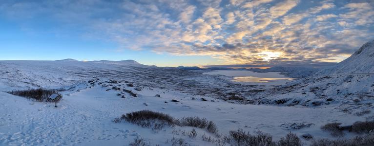 Morning view of a snowy landscape with a frozen lake. The sky is blue and the sun is rising behind clouds.
