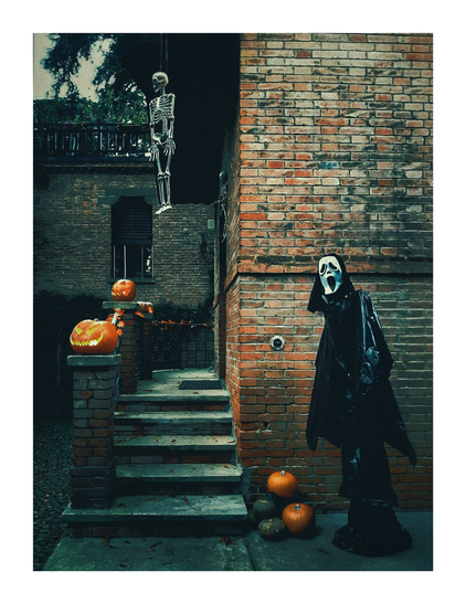 A photo of a spooky Halloween scene doorway. There is a mannequin dressed as Scream  with a white mask and black cloak on the right hand side. It stands on a little bit of paving beside a brick building. On the left above the stairs to the door there is a hanging skeleton. Three carved pumpkins sit on the steps and ground. The background includes a dark, old brick wall and a small tree.