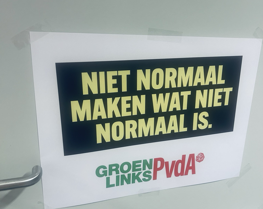 Je ziet een foto van een witte deur met een zilveren klink. Alleen het midden van de deur is in beeld. Op de deur een witte poster met een zwart vlak. In het zwarte vlak staat in gele letters ‘niet normaal maken wat niet normaal is’. Onder het zwarte vlak staat in het midden het GroenLinks-PvdA logo in de kleuren groen en rood. In groene letters staat er groen links en in rode letters staat er PvdA.