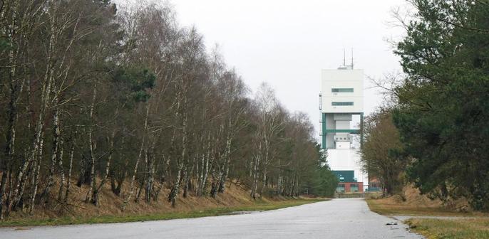Lange Landstraße führt auf einen hohen, weißen Sendemast im Wald, trüber Himmel.