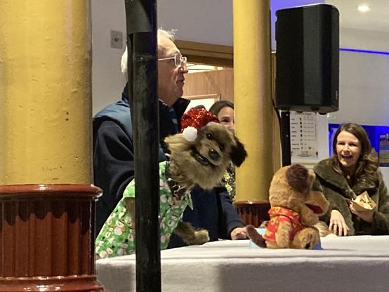 A dog puppet stares right at the camera, people around laughing, except the person who the dog is interviewing. He has no fun at all!