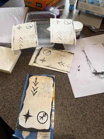An arrangement of several pieces of paper with printed designs and markings, including arrows and stars. The papers are placed on a countertop with various art supplies, including a container and tubes.