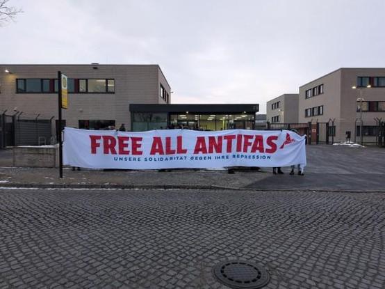 Im Hintergrund ein bunkerartiges, zweistöckiges Gebäude. Graue Ziegelsteine, einige schmale Fenster. Davor stehen Menschen hinter einem Banne. aus dem mehrere Meter langen weißen Banner steht in Großbuchstaben und in roter Farbe "Free all Antifas".
Der Boden im Vordergrund ist gepflastert und es ist ein Gullydeckel zu sehen.