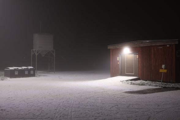 A photo of an empty parking lot at night. There is snow on the ground, a red building on the right and waste containers on the left.