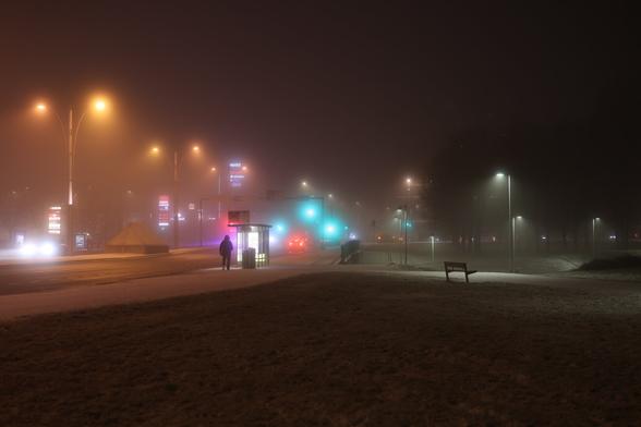 A photo of a road and street lights at night in Munkkivuori, Helsinki. There is a person walking towards a bus stop.