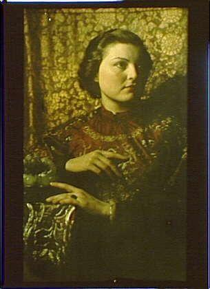 The image shows a woman sitting on a chair with her left hand resting on the armrest and holding what appears to be an ornate jade carving in her right hand. She is wearing a patterned robe with red, yellow, and gold colors, featuring intricate designs around the neckline and sleeves. The background consists of a richly textured tapestry or wallpaper that has shades of green, brown, and beige, adding depth to the overall composition. The photograph exhibits an antique look typical of early 20th-century photography, suggesting it was taken between 1906 and 1942 by Arnold Genthe (born in Austria-Hungary but later based primarily in San Francisco).