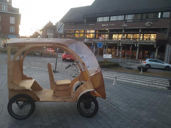 une photo du Shifti prototype V1, véhicule léger à 4 roues de cycle et une structure en bois aux formes arrondies avec un siège avant et une banquette arrière, recouverte partiellement de matière plastique, sur un trottoir au soleil couchant, au bord d'un rond point et en face d'un bâtiment moderne de plus de 3 étages, avec une allée d'arcades en béton et arborant le nom "Waux Hall" sur la façade.