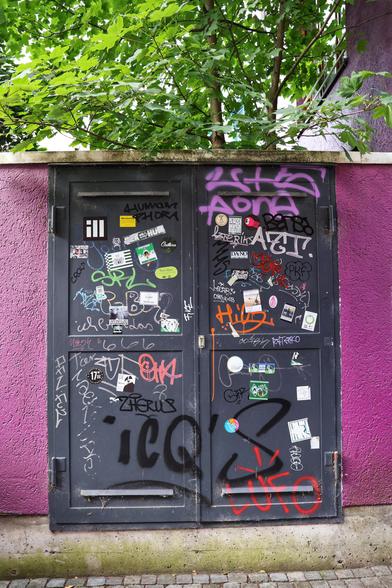 A pair of grey metal utility doors set into a purple-painted wall, heavily adorned with an eclectic mix of graffiti, stickers, and tags. The graffiti features a variety of styles, including bold lettering, abstract shapes, and colourful spray-painted designs in shades of pink, red, black, and white. Above the doors, lush green foliage from a tree adds a natural contrast to the urban, vibrant street art.