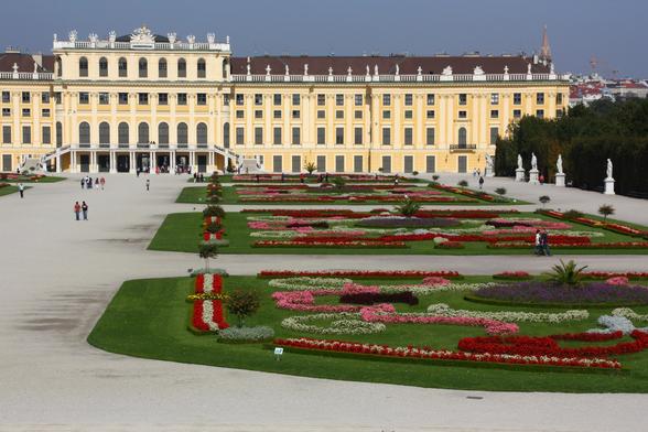 A partial view (half) of the Schönbrunn Palace and its garden. The people look like specks, showing the scale of the Palace and garden. The Palace is a pale yellow, and the grounds have wide walkways between decorative gardens with flowers and plants arranged in patterns on green grass. The sky is blue. Vienna, Austria