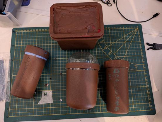 Four brown, rustic-looking, cylindrical and rectangular containers, possibly geocache hides, lying on a green cutting mat. One cylinder has a clear, rounded top.