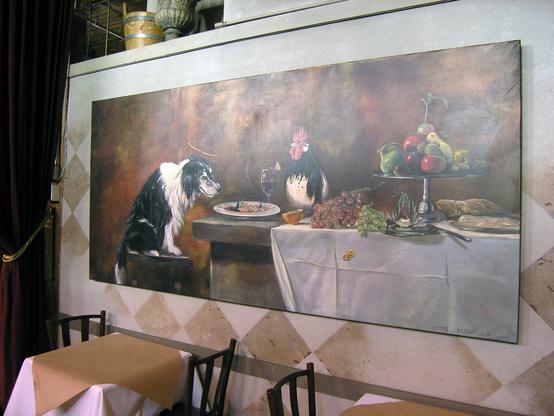 A large painting hangs on the wall inside The Pink Door restaurant in Seattle. The painting is of a feast, done in the style of Renaissance still life paintings, but this has a humorous twist. There is a large black and white dog sitting on a stool at the table, and he has a halo over his head. A rooster is also the table next to the dog. On the table are grapes, fruits and vegetables and other food, all set on a crisp white tablecloth. Beneath the painting are tables and chairs for the restaurant. The Pink Door, Seattle, Washington
