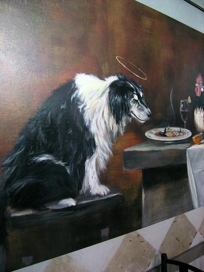 This is a closeup of the black and white dog with a halo seen in the feast painting hanging inside The Pink Door restaurant in Seattle, Washington. The dog is sitting on a stool and looking at a plate of food in front of him. There is a champagne flute glass next to the plate, and there is a glimpse of the rooster standing on the table.