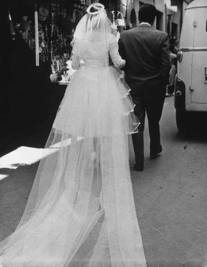 The black-and-white photograph captures a moment from what appears to be the aftermath of a wedding. The focus is on two individuals walking away: one in bridal attire with an elaborate, multi-layered veil and dress that extends down into the street, and another dressed in formal wear who might represent a groom or close relative. They are moving towards something out of frame; behind them stands what looks like a booth selling various items, possibly wedding-related decorations or goods. The setting is outdoors on a bustling city street with pedestrians around, as evidenced by people walking in the background. This scene could be interpreted as capturing the everyday reality and the busy environment contrasting against the intimate moment of marriage celebration depicted here.
