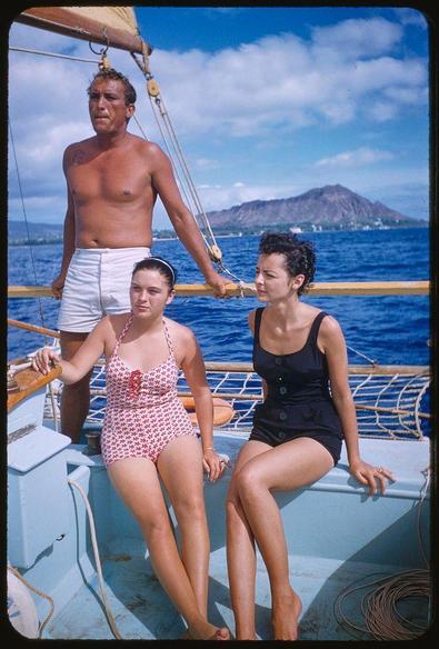 Three people on a sailboat, with the man standing and holding onto the mast while gripping some ropes. He is shirtless in white shorts, revealing his muscular arms and chest. The two women seated beside him are wearing swimsuits; one has short hair styled upwards in curlers, donning a black sleeveless dress that ends above her knees, while the other sports red-white floral patterned high-waisted one-piece with puffed shoulders and buttons down front. They look out at sea or land far off on horizon line visible behind them.

The backdrop features bright blue waters under what appears to be midday sun casting faint shadows onto boat's deck. A prominent mountain, likely a volcanic peak, rises in the distance over calm water bodies beneath it with some structures and greenery scattered around its base - indicative of an island landscape possibly Hawaii given context provided by metadata.

The colors are vivid; blues dominate due to sky and ocean while contrasts come from subjects' clothing against this backdrop. The overall scene suggests leisure activity on a sunny day, capturing the essence of beach culture or recreational sailing trip common in tropical regions like Hawaii during mid-20th century America when such photographs were taken as part journalistic assignments for Sports Illustrated magazine by Toni Frissell.