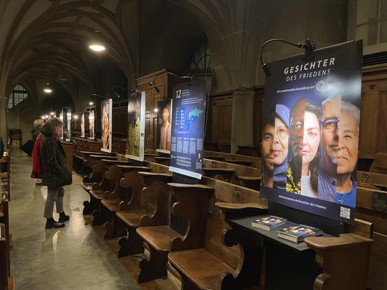 Blick in eine dunkle Kirche. An den Wänden sind Ausstellungsplakate zu sehen, die von oben angeleuchtet werden. Einige Personen stehen davor.