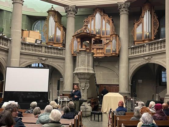 Blick in einen hellen Kirchenraum. Menschen sitzen in den Kirchenbänken und hören einem Redner zu, der mit Mikrofon vor ihnen steht.