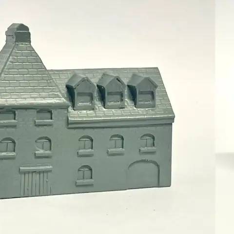 A detailed model of a building designed for 6mm scale wargames, featuring a sloped roof with three dormer windows and a textured surface that resembles slate. The facade includes several arched and rectangular windows, along with a large door at the base, all rendered in a light gray resin. The overall design suggests an industrial style, suitable for various historical or modern warfare settings.