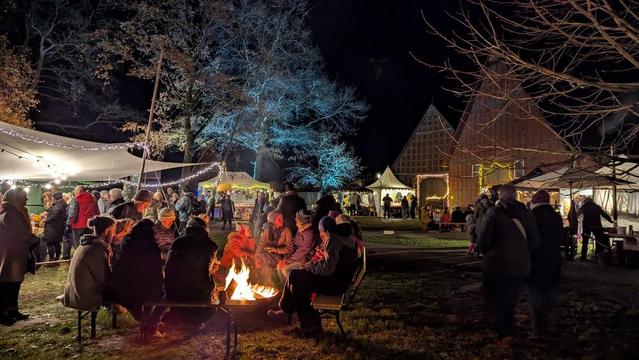 Personen sitzen um ein Lagerfeuer auf einer Wiese, im Hintergrund beleuchtete Zelte und ein Gebäude.
